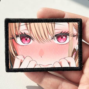 Pode incluir: Um emblema retangular bordado com uma ilustração em close de um personagem de anime com cabelo loiro, olhos rosa brilhantes e rosto corado. O personagem tem as mãos perto do queixo e o emblema possui uma borda preta espessa.