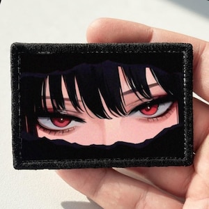 Puede incluir: Un parche rectangular bordado que presenta una ilustración gráfica de un par de ojos intensos y afilados con iris de color rojo brillante y flequillo negro en la parte superior, sobre un fondo negro con un efecto de papel rasgado.