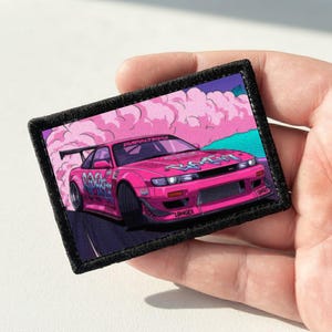 May include: A rectangular embroidered patch featuring a vibrant pink sports car drifting on a dark road. The car is surrounded by stylized pink smoke clouds against a teal background. The design includes graffiti-style text on the car body.