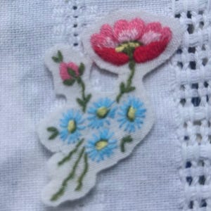 May include: A white fabric patch featuring an embroidered floral design. The embroidery includes a large pink and red flower at the top, a small pink flower bud, and a cluster of five light blue flowers with yellow centers, all connected by green stems.