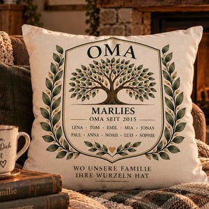 Op de afbeelding: Een vierkant gebroken wit sierkussen met een centrale boomillustratie omlijst door groene laurierbladeren. De tekst luidt OMA, MARLIES, OMA SEIT 2015, een lijst met namen en WO UNSERE FAMILIE IHRE WURZELN HAT. Bedrukt op een textuurrijke stof.