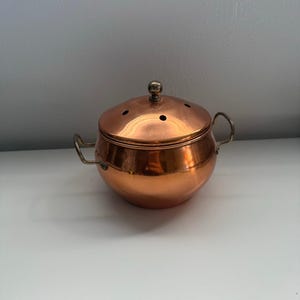 May include: A polished copper pot with a rounded body, two small side handles, and a matching lid. The lid features a central metallic knob and several small circular ventilation holes arranged in a pattern across its domed surface.