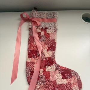 May include: A quilted Christmas stocking featuring a patchwork pattern of various red, pink, and white floral and geometric fabrics. The top cuff is trimmed with white lace and a long, light pink satin ribbon tied into a bow on the left side.