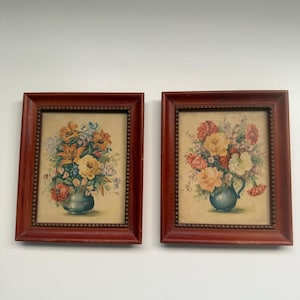 May include: Two rectangular wall art pieces featuring floral still life paintings. Each print displays a colorful bouquet of flowers in a vase, encased in a dark reddish-brown wooden frame with a beaded inner edge detail and a cream-colored mat.