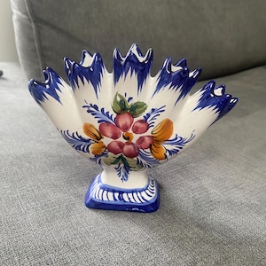 May include: A white ceramic fan-shaped vase with a scalloped top edge and a square base. The piece features hand-painted blue brushwork along the edges and a central floral motif in shades of red, orange, and green on the front surface.
