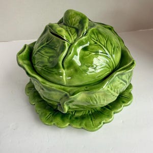 Pode incluir: Uma terrina de cerâmica verde brilhante em forma de cabeça de repolho. A peça consiste numa taça funda com uma tampa a condizer que apresenta texturas de folhas esculpidas e sobrepostas, com uma pega central em forma de folha.
