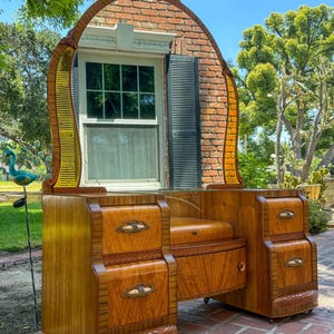 May include: A wooden vanity table featuring a large arched mirror, two side cabinets with four drawers, and decorative metal pulls. The wood grain is prominent, and the piece includes a central seat area with a small storage compartment underneath.