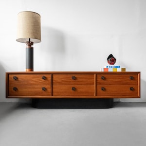 Op de afbeelding: Een lang houten dressoir met zes lades voorzien van donkere vierkante knoppen, rustend op een zwarte basis. Links staat een hoge lamp met een zwarte cilindrische voet en een beige stoffen kap. Rechts ligt een rood object op een stapel boeken.