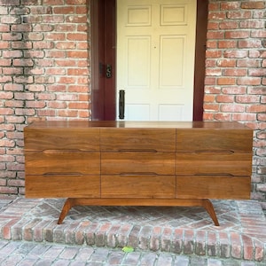 Puede incluir: Una cómoda rectangular de madera con nueve cajones dispuestos en tres columnas verticales. La pieza tiene un acabado de veta de madera marrón cálida, tiradores horizontales integrados y se apoya sobre cuatro patas de madera inclinadas.