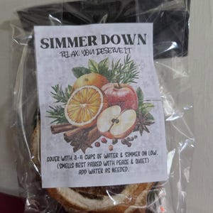 May include: A clear plastic bag containing dried fruit and spices with a label reading SIMMER DOWN RELAX. YOU DESERVE IT. The label features illustrations of orange slices, apples, cinnamon sticks, and rosemary, with instructions to cover with 3-4 cups of water.