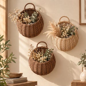 Könnte beinhalten: Drei runde, geflochtene braune Weidenkörbe zur Wandmontage, jeweils mit einem stabilen, bogenförmigen Griff an der Oberseite. Die Körbe hängen an der Wand und sind mit Trockenblumenarrangements aus weißen Blüten und Eukalyptusblättern gefüllt.