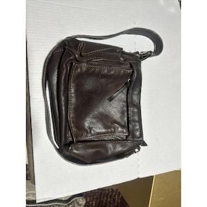 May include: A dark brown leather shoulder bag featuring a textured surface, a single adjustable strap, and a prominent square front pocket with visible stitching details. The bag has a rounded silhouette and a zipper closure on the main compartment.