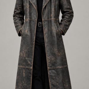 May include: A long, dark brown distressed leather trench coat featuring a wide lapel collar, long sleeves, and two side pockets. The coat has a weathered, textured finish with visible seam detailing along the front and hemline, worn over a black shirt.