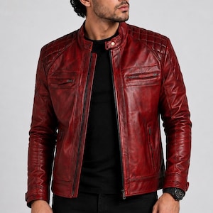 May include: A deep red leather jacket featuring a mandarin collar with a snap button closure, a full-length front zip, and quilted shoulder panels. The jacket includes two horizontal zipped chest pockets and two lower side pockets with zips.