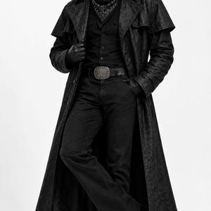 May include: A black long duster coat worn over a black button-down shirt and black trousers. The outfit includes a black wide-brimmed hat, a patterned black neckerchief, a black leather belt with a large silver buckle, and black leather gloves.