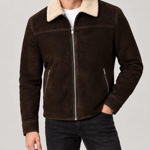 Puede incluir: Una chaqueta de ante marrón oscuro con cuello de piel sintética color crema, cierre de cremallera metálica plateada y dos bolsillos laterales con cremalleras. La chaqueta se lleva sobre una camiseta blanca lisa y unos vaqueros negros.