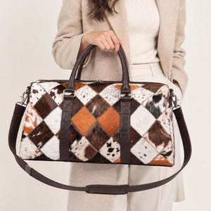 May include: A duffel bag featuring a patchwork design of cowhide in shades of brown, white, and black. The bag has dark brown leather handles, a matching detachable shoulder strap, and a top zipper closure with silver-toned metal hardware.