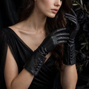 May include: A pair of black leather gloves featuring a wide cuff design adorned with four matching black buttons on the outer wrist area. The gloves have a smooth finish and extend past the wrist, providing a fitted appearance on the hands.