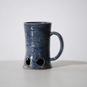 Könnte beinhalten: Ein hoher, zylindrischer Keramikbecher mit einer tiefblauen, gesprenkelten Glasur und einer strukturierten, geriffelten Oberfläche. Der Boden weist zwei unregelmäßige, kreisförmige Ausschnitte auf und ein geschwungener Henkel ist seitlich angebracht.