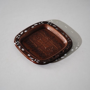 May include: A square copper-toned metal tray featuring a hammered texture on the inner surface and an intricate openwork border. The centre displays a relief design of two classical columns with ornate capitals connected by horizontal bars.