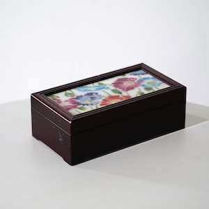 May include: A rectangular dark brown wooden box featuring a hinged lid with an inset ceramic tile. The tile displays a colorful floral pattern with painted red, blue, and purple poppy-like flowers against a white background.