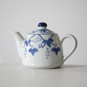 May include: A white ceramic teapot featuring a hand-painted blue pattern of grape clusters and vine leaves. The teapot includes a matching lid with a solid blue knob, a curved handle, and a tapered spout, all decorated with a delicate blue vine motif.