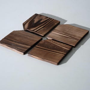 May include: A set of four square wooden coasters featuring a dark brown stained finish with visible wood grain patterns. Each coaster has a flat surface and straight edges, displaying a natural texture and deep, rich colour tones throughout the wood.