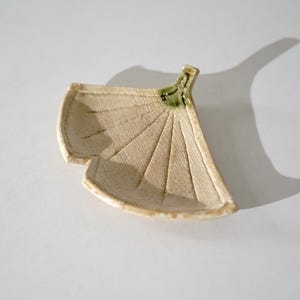 Pode incluir: Um pequeno prato de cerâmica em forma de leque inspirado numa folha de ginkgo. A peça apresenta um acabamento de esmalte craquelado de cor creme com veios texturizados e um acento verde distinto na base do caule.