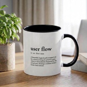 May include: A white ceramic mug with a black interior and handle, featuring the printed dictionary-style definition of user flow: A beautiful, logical path mapped out for a human being to achieve a goal, which is immediately bypassed by the actual user clicking a random broken footer link instead.