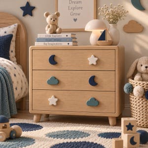 May include: A light wood three-drawer chest featuring decorative drawer pulls shaped like navy blue moons, white stars, and teal clouds. The chest stands on four short, rounded legs and has a smooth, natural wood grain finish throughout.