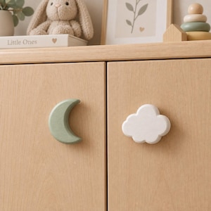 May include: Two wooden cabinet knobs attached to light-toned wood doors. One knob is shaped like a crescent moon in a muted sage green colour, and the other knob is shaped like a fluffy cloud in a solid white colour with a smooth, matte finish.