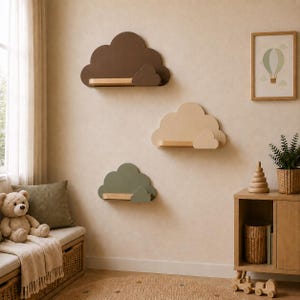 Peut inclure: Trois étagères murales en forme de nuages, chacune dotée d'une tablette en bois plate. L'étagère supérieure est marron foncé, celle du milieu est beige et l'étagère inférieure est vert sauge. Chaque nuage possède un petit accent nuageux.