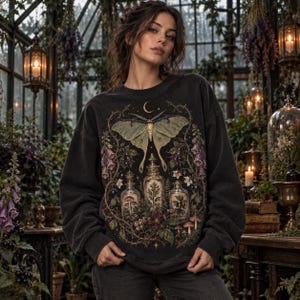 May include: A charcoal gray long-sleeve sweatshirt featuring a large graphic print of a pale green moth centered above three glass apothecary bottles filled with botanical elements, surrounded by intricate vines, flowers, and small mushrooms.