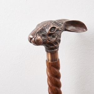 Könnte beinhalten: Ein Gehstock mit einem detaillierten, bronzefarbenen Metallgriff in Form eines Kaninchenkopfes mit langen Ohren. Der Griff ist an einem dunkelbraunen Holzschaft befestigt, der eine markante, gedrehte und spiralförmig geschnitzte Textur aufweist.