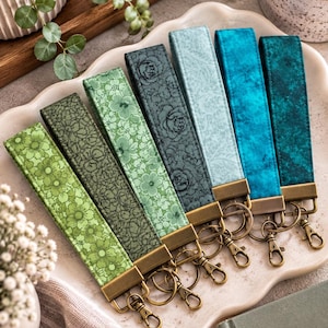 May include: A set of six fabric wristlet keychains featuring various shades of green and blue. Each keychain has a floral or textured pattern, a bronze-toned metal clasp, and a split ring. The wristlets are arranged in a row on a scalloped tray.