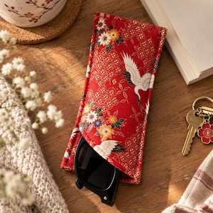 Puede incluir: Un estuche rectangular de tela para gafas con fondo rojo vibrante, un patrón geométrico dorado repetitivo, ilustraciones de grullas blancas y detalles florales coloridos. El estuche contiene unas gafas de sol negras parcialmente visibles.