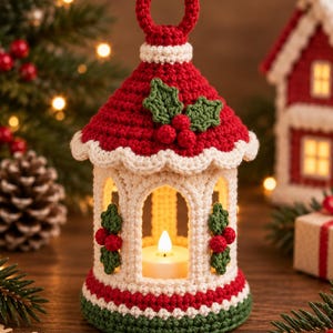 May include: A crocheted lantern in red, white, and green featuring a pointed roof with a hanging loop, arched openings, and decorative holly leaf and berry appliqués. A small white battery-operated tea light candle sits inside the structure.