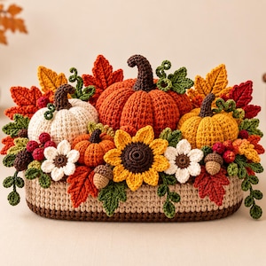Op de afbeelding: Een gehaakt tafelstuk met drie pompoenen in wit, oranje en geel, geaccentueerd met een centrale zonnebloem, witte bloemen, rode bessen, bruine eikels en diverse herfstkleurige bladeren, gerangschikt in een beige en bruine ovale mand.