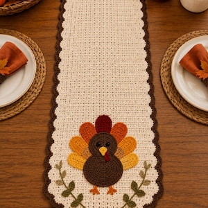May include: A cream-colored crocheted table runner with a dark brown scalloped border. The runner features a central applique of a brown turkey with a multi-colored tail in shades of red, orange, and yellow, flanked by two green leafy branches.