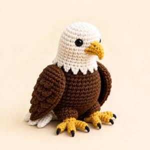 May include: A crocheted bald eagle soft toy featuring a white head, dark brown body and wings, a bright yellow beak, and yellow feet with black-tipped claws. The toy has small black plastic eyes and a textured, yarn-based construction.