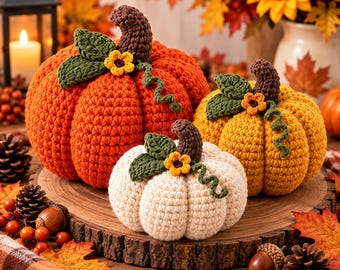 May include: Three crocheted pumpkins in orange, yellow, and cream, each featuring a dark brown stem, green leaves, a small yellow flower, and a green curly vine. The pumpkins are arranged on a round wooden slice with a textured bark edge.