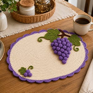May include: An oval crochet placemat in cream with a scalloped purple border. The placemat features a large, textured cluster of purple grapes with green leaves and vines, and a smaller matching grape cluster motif in the lower left corner.