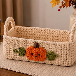 May include: A rectangular cream-colored crochet basket with two handles, featuring a central orange pumpkin appliqué with a brown stem, black bead eyes, a stitched smile, and two green leafy accents on either side of the pumpkin.