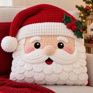 May include: A square crochet pillow shaped like a Santa Claus face. The pillow features a red hat with a white pom-pom, a white brim, a peach-colored face with black eyes, pink cheeks, a white mustache, and a white beard. Green holly leaves and red berries are attached to the hat.
