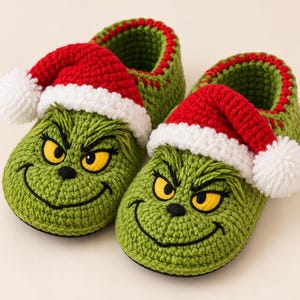 Peut inclure: Une paire de chaussons en crochet vert représentant un visage de personnage aux yeux jaunes et au sourire noir brodé. Chaque chausson est surmonté d'un bonnet de Père Noël en crochet rouge et blanc avec un pompon blanc sur le côté.