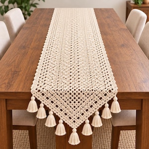 Puede incluir: Un camino de mesa largo y rectangular de color crema con un intrincado patrón de nudos estilo macramé con un diseño de rejilla de diamantes. El camino está rematado con múltiples borlas a juego que cuelgan de los bordes puntiagudos.