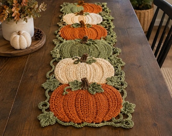 May include: A crochet table runner featuring a repeating pattern of pumpkins in shades of orange, cream, and olive green. The design includes interconnected crocheted leaves and vines, with each pumpkin topped by a small brown stem.