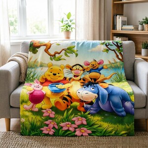 May include: A soft fleece throw blanket featuring a colourful illustration of Winnie the Pooh, Tigger, Piglet, Eeyore, and Roo in a grassy meadow with pink flowers. The characters are positioned near a blue pond under a tree with green leaves.