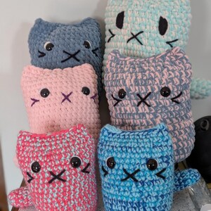 May include: A collection of six handmade crochet cat plushies in various colors including blue, pink, and variegated yarn. Each soft toy features embroidered facial details, including black eyes and a stitched mouth, with pointed ears.