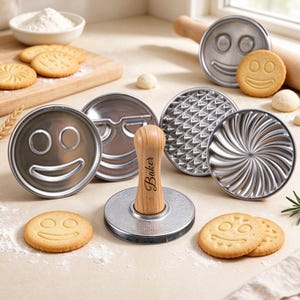 Könnte beinhalten: Ein Set aus runden Metall-Keksstempeln mit verschiedenen Gesichtern und Mustern, inklusive eines Holzgriffs mit der Gravur Baker. Die Stempel sind zusammen mit gebackenen Keksen angeordnet, die passende Abdrücke auf einer hellen Fläche zeigen.